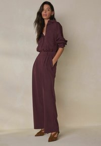 Oversize skjorte i mørkerød med knapdetaljer, kombineret med vidde bukser med plisseringer og lommer. Brune sko med spidse snuder fuldender outfittet.