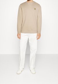 Sudadera beige con puños acanalados, que presenta un pequeño logo bordado en amarillo. Combinada con pantalones rectos blancos y zapatillas blancas.