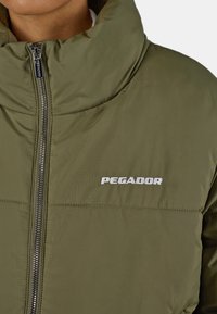 Olivově zelená péřová bunda s vysokým límcem, vybavená stříbrným zipem a bílým logem "PEGADOR" na hrudi. Hladká, lehká látka.