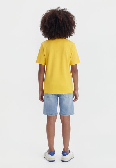 Niño con cabello rizado, vestido con una camiseta amarilla, pantalones cortos de mezclilla azul claro y zapatillas blancas, de pie de espaldas sobre un fondo liso.