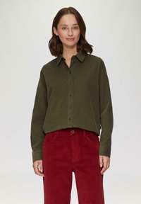 Chemise en velours côtelé vert olive avec boutons, manches longues et col, associée à un pantalon en velours côtelé rouge avec poches avant.