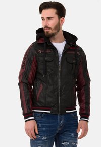 Schwarz und kastanienbraune Kapuzenjacke mit gestepptem Muster, mehreren Reißverschlusstaschen und gestreiften Bündchen. Getragen über einem weißen T-Shirt und Jeans.