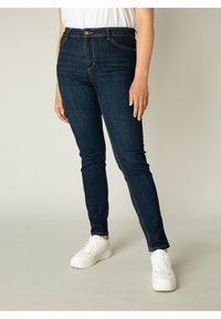 Donkere denim skinny jeans met een hoge tailleband, voorzien van stikseldetails en een knoopsluiting aan de voorkant, gecombineerd met witte sneakers.