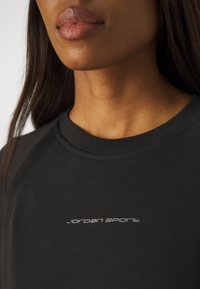 Gros plan sur le bas du visage et le cou d'une femme portant un t-shirt noir à col rond avec l'inscription "Jordan Sport" en blanc sur la poitrine.