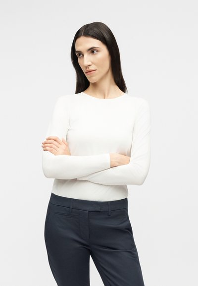 Γυναίκα με μακριά σκούρα μαλλιά, φορώντας λευκό μακρυμάνικο μπλουζάκι και σκούρο παντελόνι, στέκεται με σταυρωμένα τα χέρια, κοιτάζοντας προς το πλάι μπροστά σε απλό φόντο.