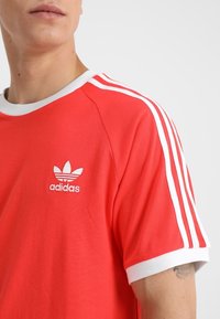 Punainen puuvillainen t-paita, jossa valkoiset yksityiskohdat, pyöreä pääntie ja kolme valkoista raitaa olkapäillä. Adidas-logo kirjailtuna rintakehään.
