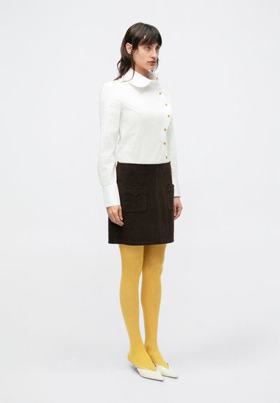 Biela košeľa na zapínanie s veľkým golierom, hnedá semišová sukňa s dvoma prednými vreckami, žlté pančuchy a biele lodičky so špicatou špičkou.