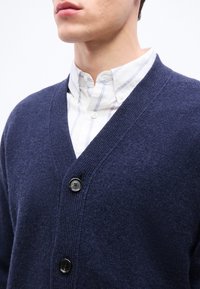 Άνδρας που φοράει λευκό πουκάμισο με μπλε ρίγες κάτω από μπλε ναυτικό καζάκα με κουμπιά.