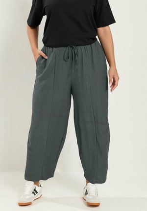 Persona que lleva pantalones sueltos de color verde oscuro con cordón, camiseta negra, zapatillas blancas con rayas negras, de pie con una mano en el bolsillo.