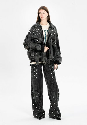 Joven mujer que lleva una chaqueta de mezclilla negra de gran tamaño y pantalones de pierna ancha con tachuelas de puntos blancos, combinados con una camiseta gráfica y zapatos negros.