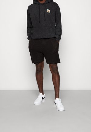 Osoba ubrana w czarną bluzę z kapturem z nadrukiem, czarne szorty i białe buty Nike, stojąca z rękami w kieszeniach na jednolitym tle.