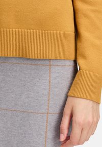 Kéz pihen a szürke szoknya mellett, amely finom barna kockás vonalakkal díszítve, mustárszínű bordázott pulóver ujjaival és aljával.