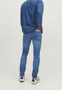 Džínová košile tmavě modré barvy s knoflíkovým límečkem, kombinovaná se světle modrými džínami s prošíváním na zadních kapsách a bílými teniskami.