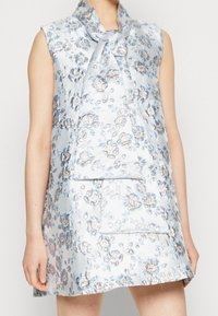 Vaaleansininen ilman hihoja oleva mekko kukkakuosisena, jossa on kimaltavia yksityiskohtia. Mekossa on leveä kaulus ja suorakulmainen tasku edessä.