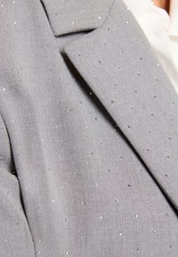 Blazer gris con tejido texturizado y pequeños adornos brillantes dispersos por toda su superficie. Solapas estructuradas que añaden detalle.