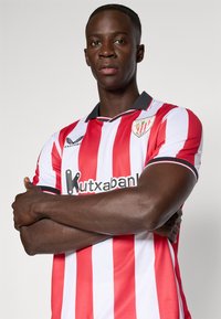 Männlicher Athlet mit verschränkten Armen, der ein rot-weiß gestreiftes Trikot des Athletic Club Bilbao mit Sponsor-Logos trägt.