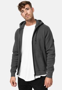 Sweat à capuche gris avec fermeture éclair, cordon de serrage, poches avant et manches côtelées. Porté sur un T-shirt blanc, avec une coupe décontractée.