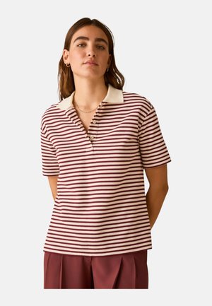 Donna che indossa una polo a maniche corte crema e bordeaux a righe, con colletto crema e pantaloni bordeaux, in piedi con le mani dietro la schiena.