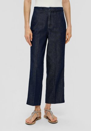 Personne portant un jean large bleu foncé raccourci, associé à des sandales plates argentées à brides, debout sur un fond uni.