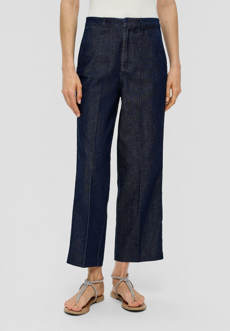 Personne portant un jean large bleu foncé raccourci, associé à des sandales plates argentées à brides, debout sur un fond uni.