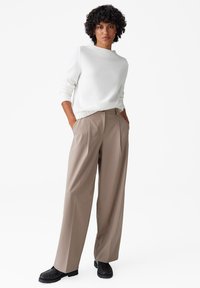 Witte gebreide trui met structuur, gecombineerd met beige hoog-waisted wijde broek en zwarte instappers. Het model staat met de handen in de zakken.