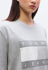 Γκρι φούτερ με έντονο γραφικό σχέδιο που περιλαμβάνει το κείμενο "TOMMY JEANS", ορθογώνιο μοτίβο και απαλό, υφασμάτινο υλικό.