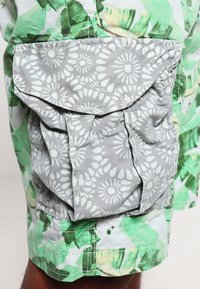 Zielone szorty w kwiatowy wzór mają szary materiałowy kieszeń z kwiatowym motywem, lekko marszczoną dla nadania tekstury i praktyczności w designie.