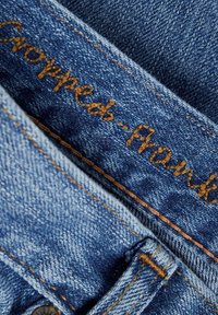 Jeans in denim accorciati con un colore blu sbiadito, caratterizzati da cuciture dorate e un'etichetta ricamata con la scritta "Cropped-Front."