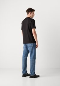T-shirt noir à manches courtes en coton, associé à un jean en denim bleu. Modèle vu de dos, portant des chaussures noires et ayant des cheveux courts et bouclés.