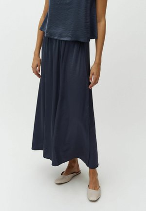 Femme portant un haut sans manches bleu marine et un pantalon large assorti, avec des chaussures beiges sans lacets, debout contre un fond uni.