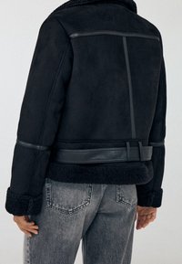 Persoon in een zwarte schapenvachtjas met leren randen en manchet details, gecombineerd met vervaagde grijze jeans, getoond vanaf de achterkant.