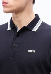 Homme portant un polo bleu marine avec un col rayé blanc et le logo "BOSS" brodé sur la poitrine, tourné vers la gauche, cadré au niveau du cou.