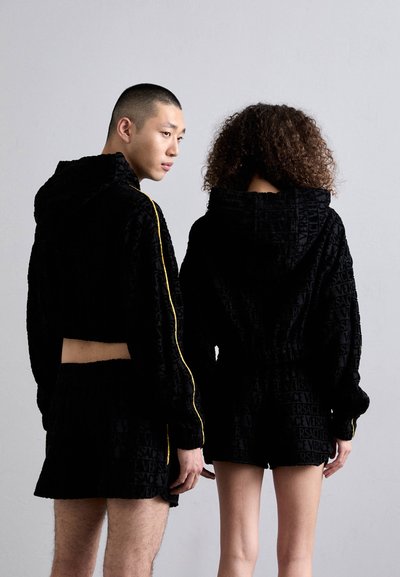 Jachetele scurte cu glugă de culoare neagră și pantaloni scurți au o textură catifelată, cu logo-ul Versace embosat, accentuate de o dâră galbenă pe jachetă.