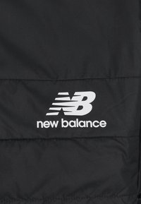 Svart jacka med quiltad textur, med en framträdande vit New Balance-logotyp och bokstäver på nedre högra sidan.