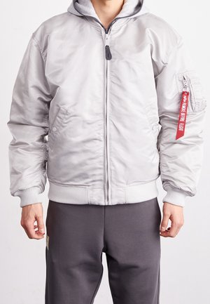 Man wearing light gray bomber jacket with front zipper and red "Remove Before Flight" tag on left sleeve pocket, paired with dark gray pants.