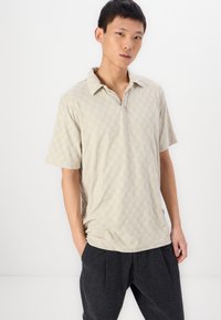 Polo shirt scurt, de culoare bej deschis, cu un model texturat în carouri și guler, asortat cu pantaloni texturați de culoare gri închis.