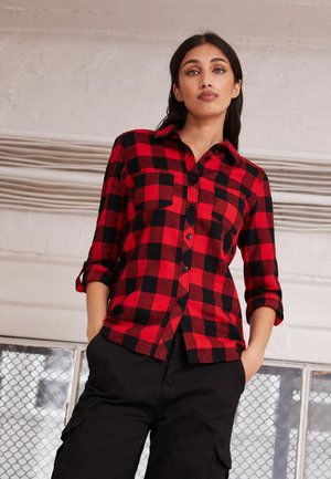 Chemise en flanelle à carreaux rouges et noirs avec manches longues, fermeture à boutons et deux poches poitrine. Portée avec un pantalon cargo noir dans un environnement lumineux.