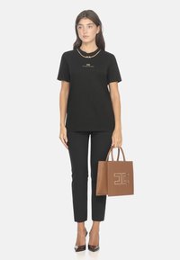 T-shirt nera in cotone con logo dorato, abbinata a pantaloni neri aderenti, borsa a tracolla in pelle marrone e tacchi alti marroni.