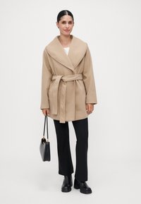 Kvinne iført en beige belted frakk, svarte bukser, svarte platåsko, og holder en svart håndveske, stående mot en ensfarget hvit bakgrunn.