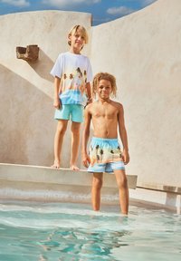 Δύο αγόρια δίπλα σε μια πισίνα; το ένα στέκεται ξυπόλητο στην άκρη φορώντας πουκάμισο και σορτς με τύπωμα φοίνικας, ενώ το άλλο βυθίζεται στο νερό φορώντας αντίστοιχα μαγιό. 