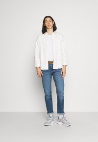 Camisa blanca de botones, camiseta blanca, vaqueros azul, cinturón marrón y zapatillas grises con acentos rojos y azules. Fondo liso.