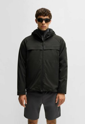 Giacca nera impermeabile con cappuccio, chiusura zip frontale e tasca sul petto. Indossata sopra pantaloni corti grigi. Tessuto liscio e vestibilità atletica.