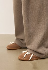 Papuci din suedă maro cu șireturi albe purtați sub pantaloni largi din lână bej, stând pe un podea netedă.