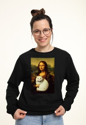 Femme souriante avec des lunettes portant un sweat-shirt noir montrant la Mona Lisa tenant un chat blanc imprimé sur le devant.