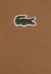 T-shirt en tissu tricoté marron avec un logo crocodile vert brodé, bordé de blanc. La texture est lisse avec un motif côtelé.