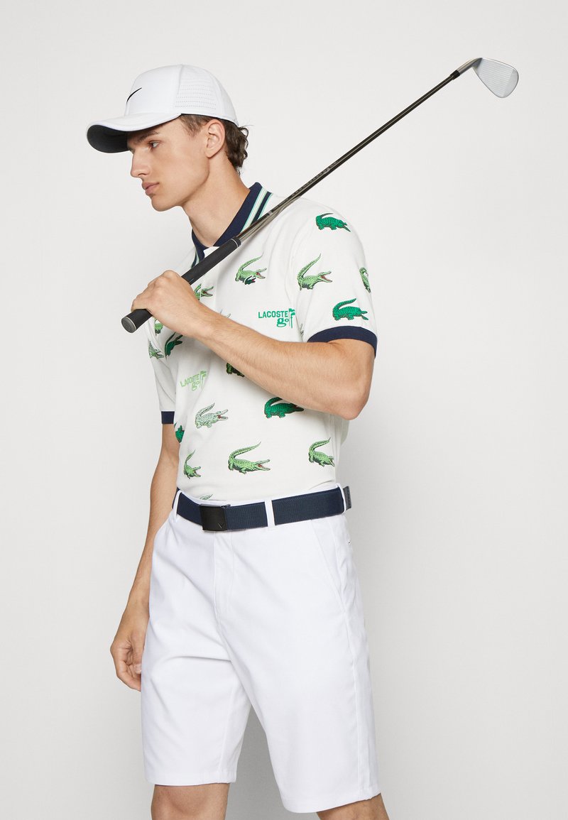 Jeune homme en tenue de golf blanche avec des logos de crocodile verts, tenant un club de golf sur son épaule, regardant sur le côté.