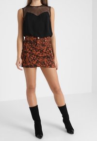 Blouse sans manches noire avec un col en mesh, associée à une mini-jupe en imprimé serpent orange et des bottes noires  à talons hauts jusqu'aux genoux.