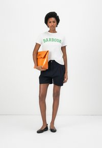 Valge T-särk rohelise "BARBOUR" tekstiga, tumesinised lühikesed püksid ja oranž klutš. Mudel kannab musti lapikesi, seistes ühtlase tausta ees.