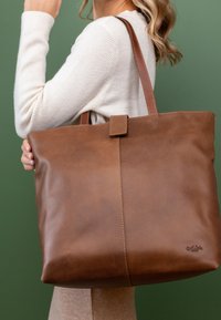 Femme en pull beige tenant un grand sac fourre-tout en cuir marron sur l'épaule, sur fond vert.