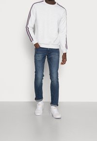 Light grey sweatshirt with navy and red striped sleeves, paired with blue jeans and white sneakers. Casual fit, ribbed neckline and cuffs.